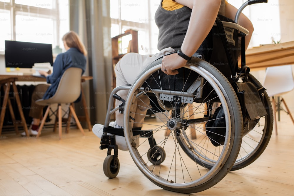 Low section of young disable female sitting in wheelchair in front of camera while moving towards colleague sitting by one of tables