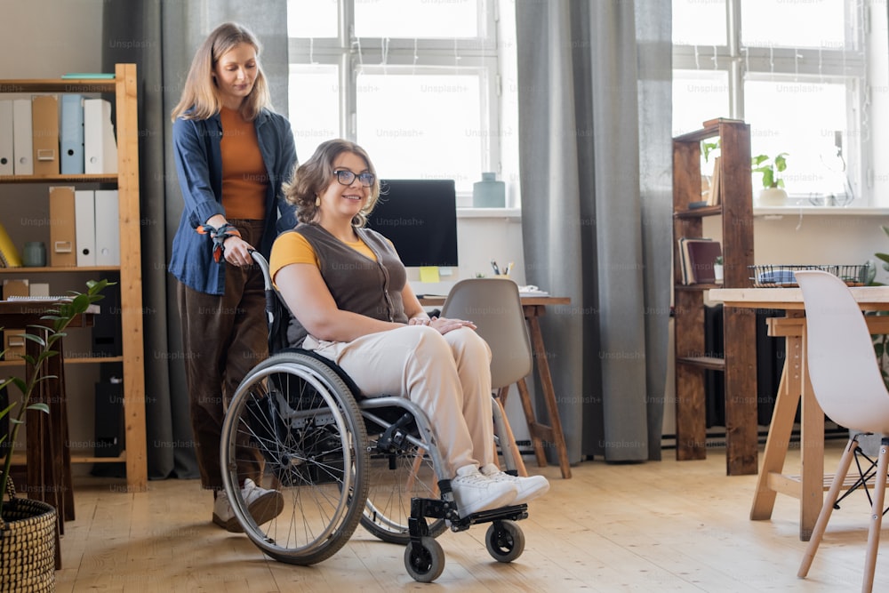 Young smiling woman in wheelchair looking forwards while her sister or friend standing behind and helping her with moving around