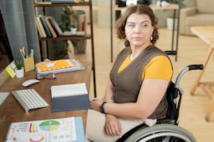 휠체어를 탄 진지하거나 생각에 잠긴 젊은 사업가가 컴퓨터 앞 책상 옆에 앉아 사무실에서 데이터 작업을 하는 동안 옆을 바라보고 있다