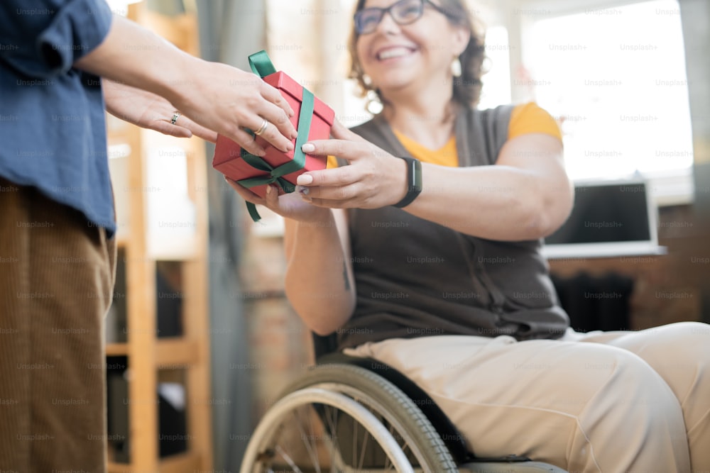 Joven alegre mujer de negocios discapacitada en silla de ruedas pasando por una caja de regalo llena con regalo de Navidad o cumpleaños a su colega femenina