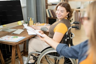 Young smiling businesswoman in wheelchair taking clipboard with financial document from hands of colleague while working with data in office