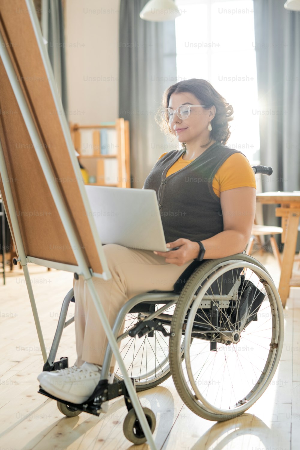 Jeune femme d’affaires handicapée regardant l’écran d’un ordinateur portable tout en préparant une présentation pour ses collègues devant un tableau blanc dans un bureau moderne