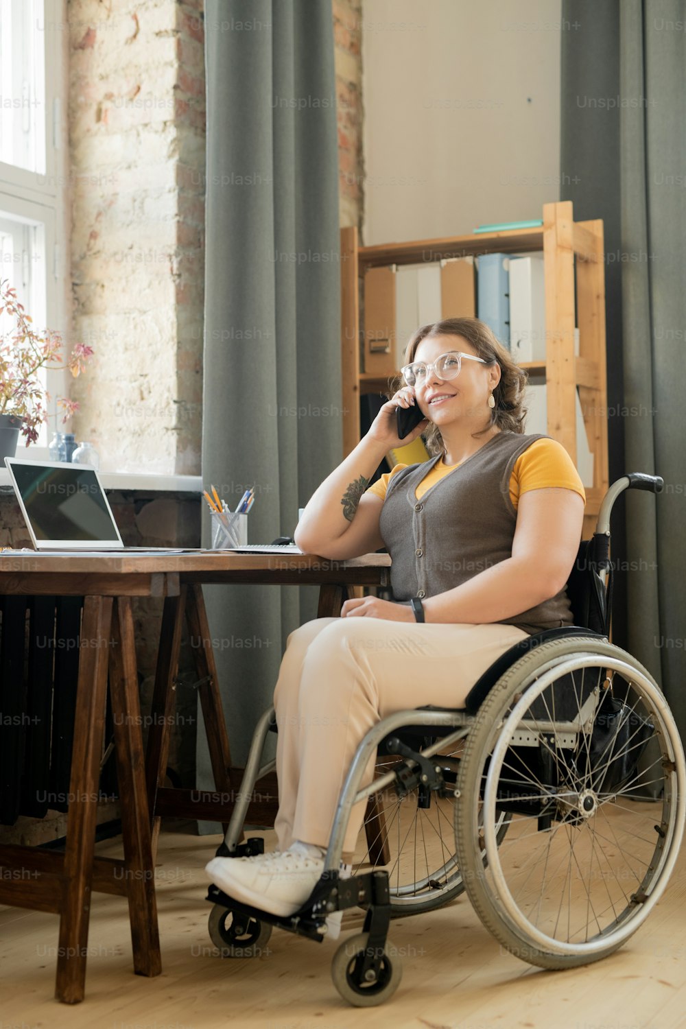 Happy young handicape une agente ou une secrétaire de bureau en fauteuil roulant assise près d’une table et consultant les clients sur téléphone portable par lieu de travail