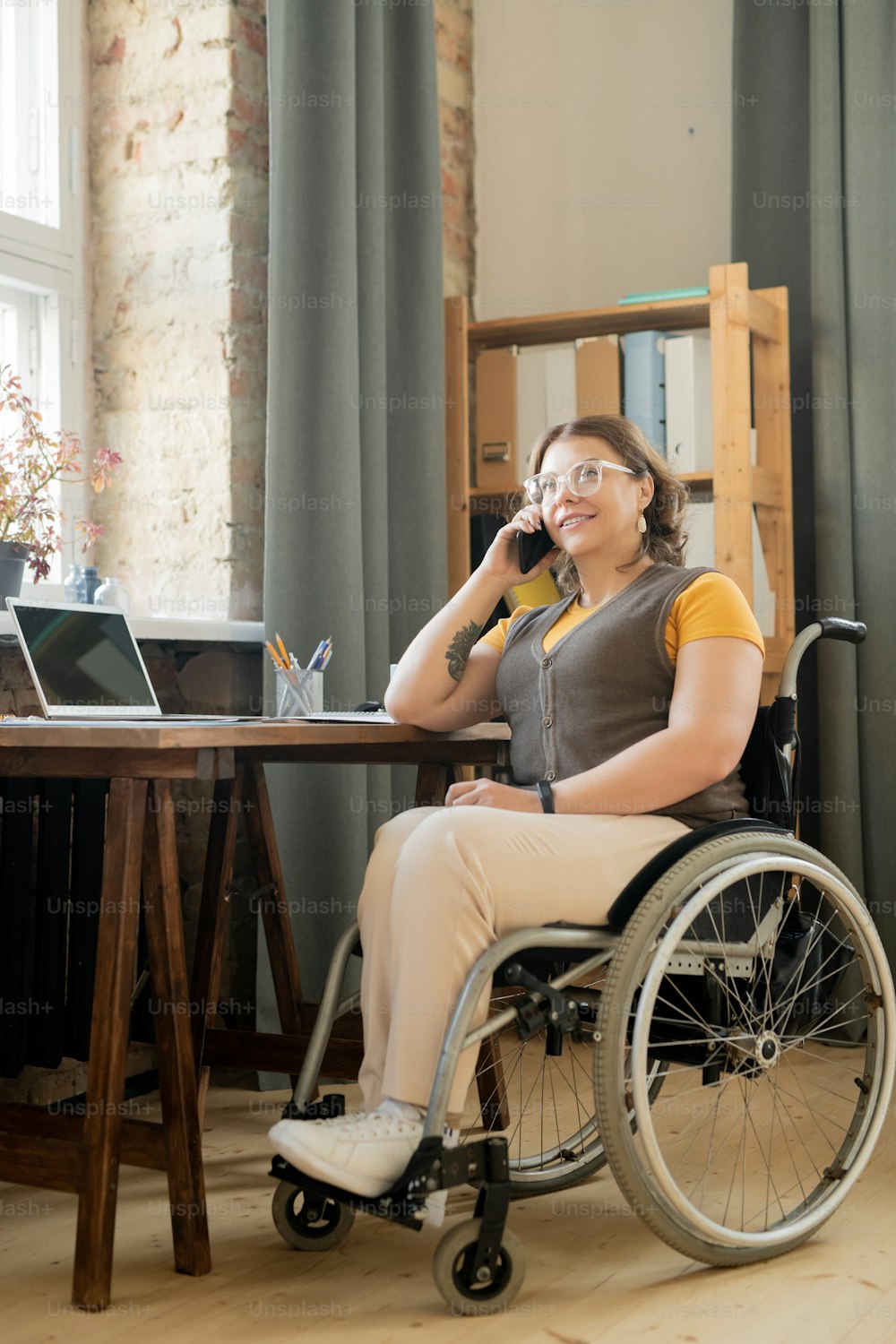 テーブルサイドに座り、職場で携帯電話でクライアントに相談する車椅子の幸せな若い障害者女性エージェントまたはオフィス秘書