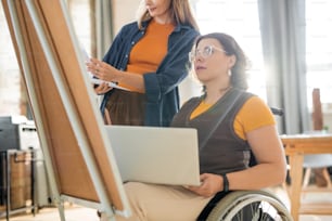 Young businesswoman in casualwear helping her disable colleague with presentation while standing in front of whiteboard in office