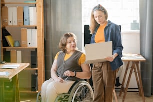 Young confident businesswoman with laptop showing information for report or business project to her disable colleague in wheelchair
