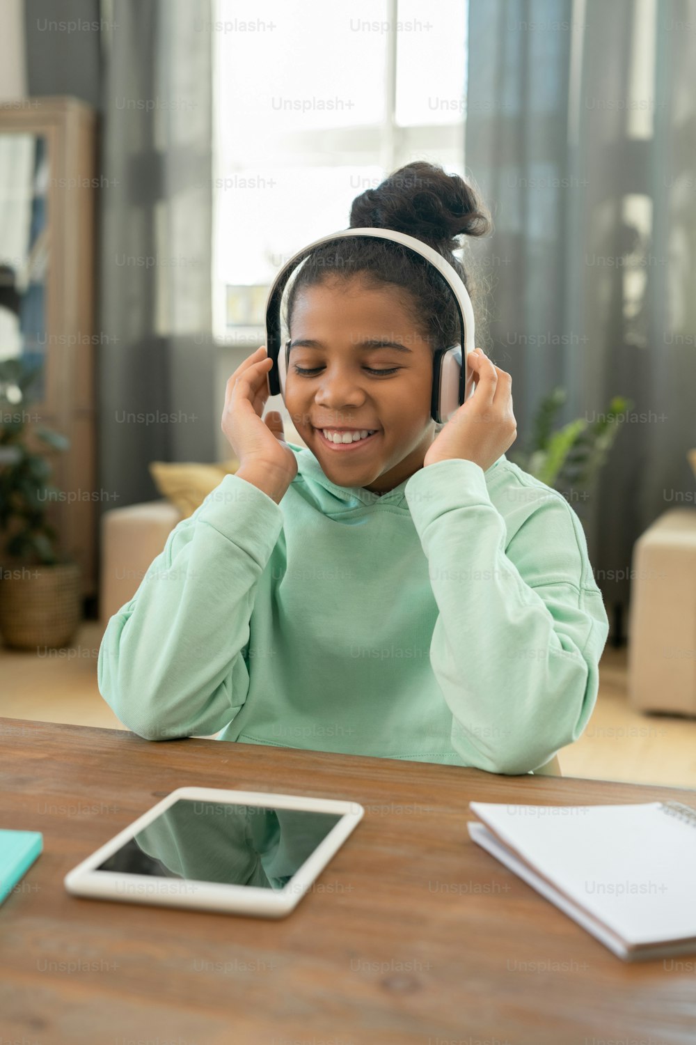 Cheerful African schoolgirl with her eyes closed sitting by table in living-room against large window and listening to joyful music in headphones