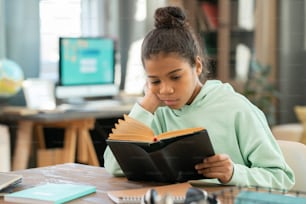 Ernstes Grundschulmädchen in Freizeitkleidung, das am Tisch im Wohnzimmer gegen den Arbeitsplatz mit Computer sitzt und Schulliteratur liest