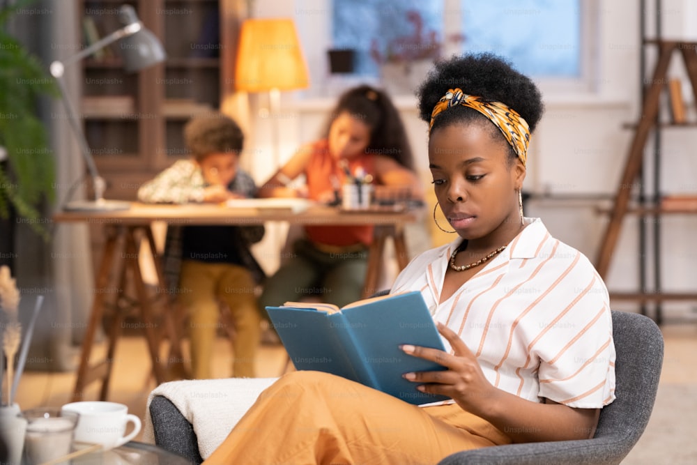 Young African woman in casualwear sitting in soft grey armchair by small table and reading book against her two children doing homework