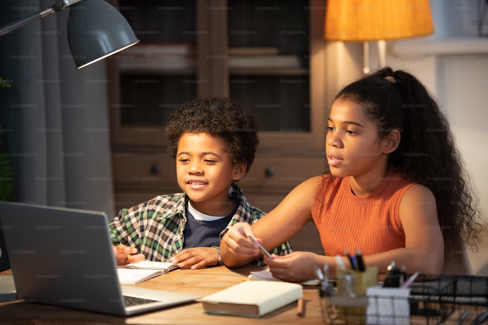 Two cute siblings of African ethnicity sitting by table in front of laptop and taking part in online lesson while listening to teacher explanation