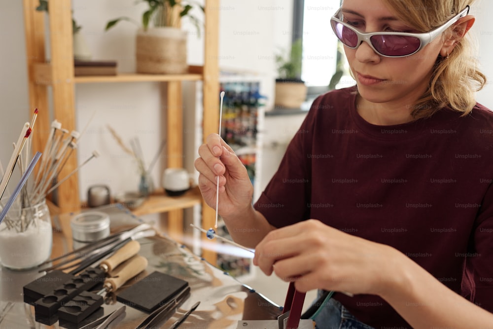 Joven artesana con gafas protectoras sentada en el taller y decorando una pieza de trabajo de vidrio en un palo mientras la sostiene sobre la mesa