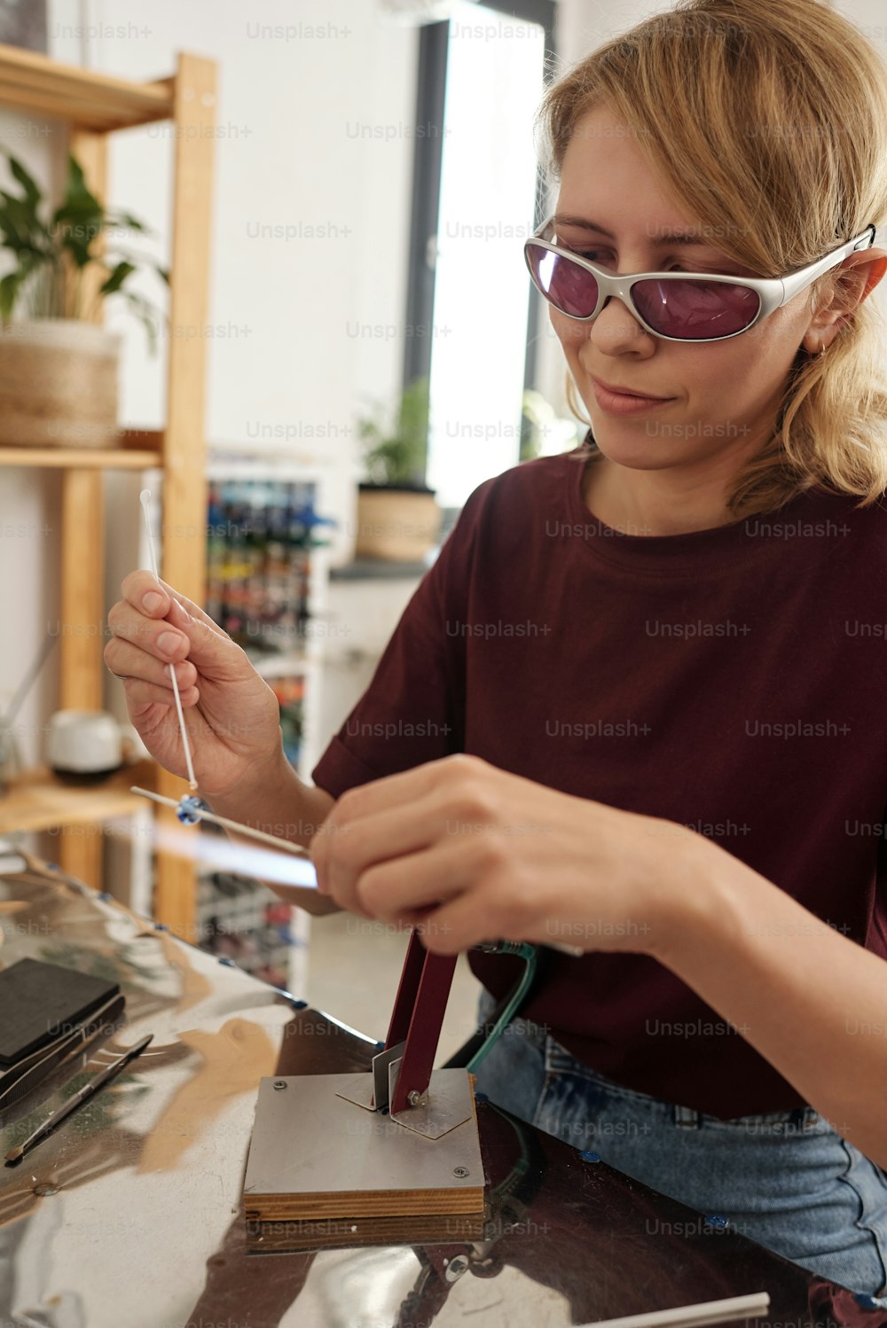 保護眼鏡とカジュアルウェアを着た若い女性職人が、ガラスのワークピースをテーブルの上にかざしながら、溶けたペンキで棒に飾ります