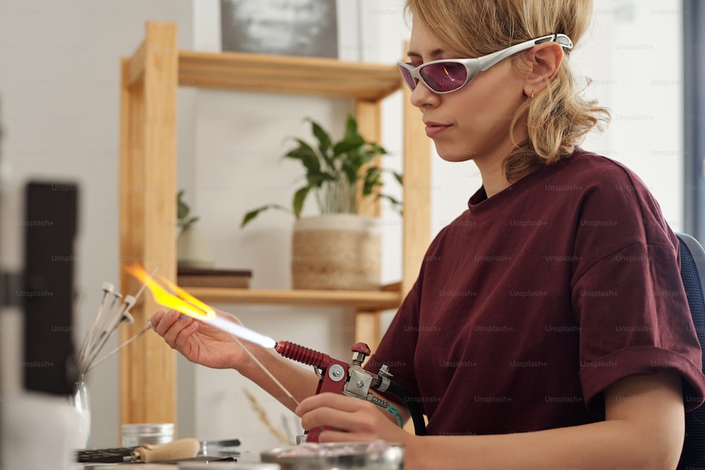 Junge ernsthafte Kunsthandwerkerin mit Schutzbrille, die in der Werkstatt sitzt und Glaswerkstück mit Feuer brennt, während sie es über den Brenner hält