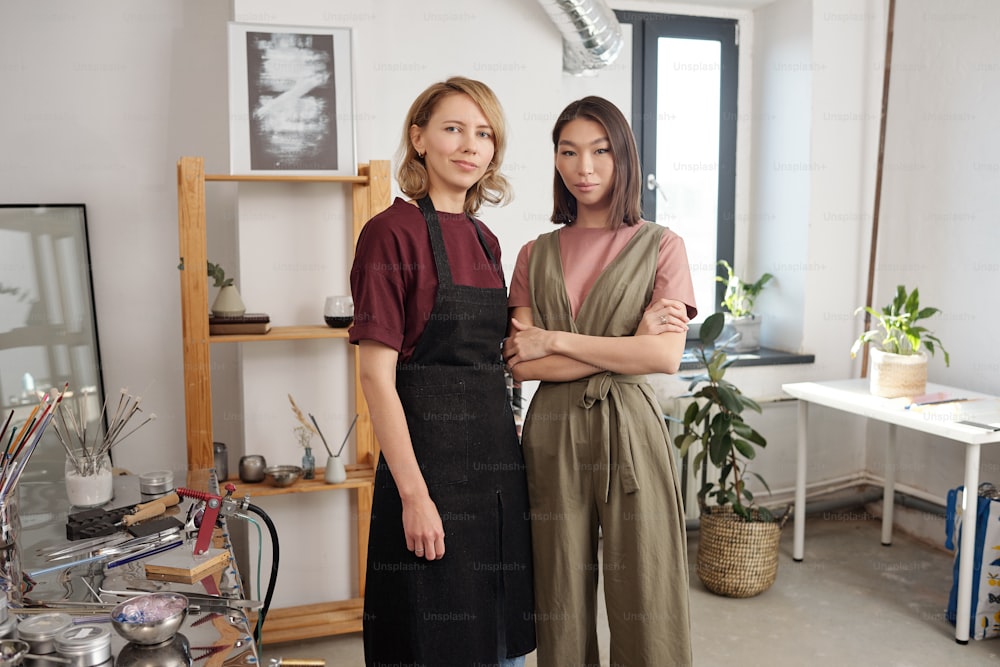 Two young intercultural female professional lampworkers in workwear standing next to one another in front of camera in large workshop
