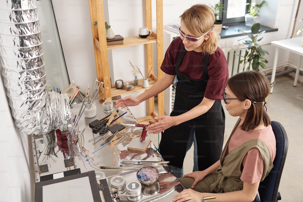 Young contemporary craftswoman in workwear and eyeglasses showing her trainee how to use burner for processing glass workpieces