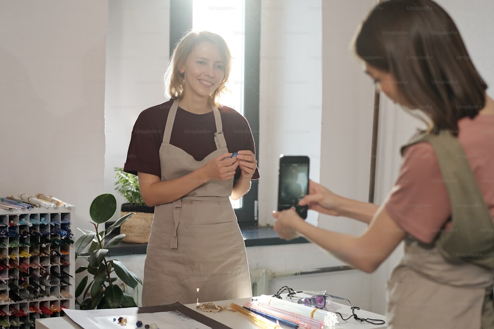 Happy young blond female with handmade glass bead standing by workplace while brunette girl with smartphone taking photo or video
