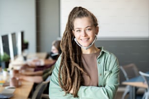 Happy young female operator with dreadlocks standing in front of camera against row of desks and computers and crossing arms by chest