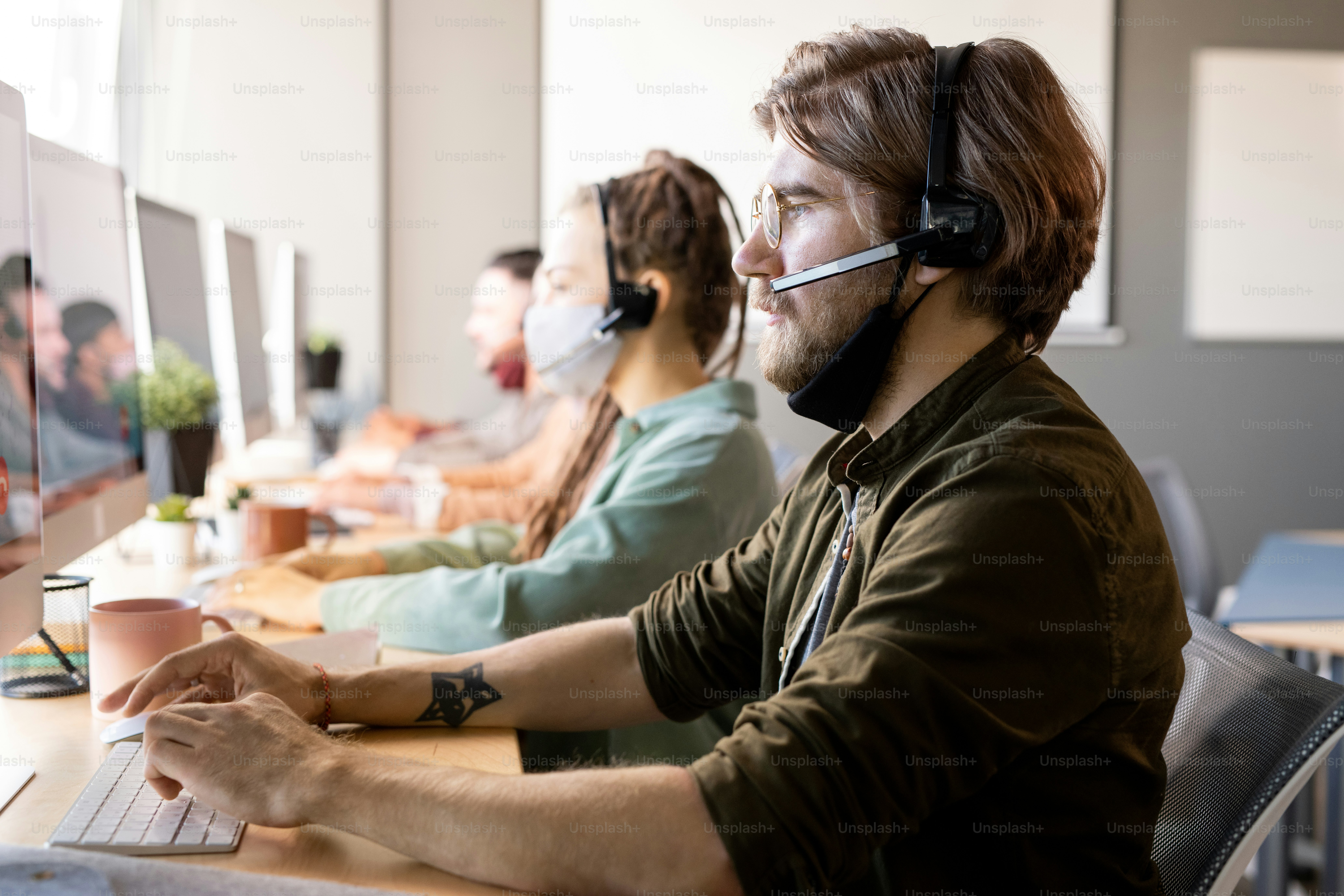 Junger, ernster, bärtiger männlicher Bediener mit Schutzmaske unter dem Kinn, der Kunden berät, während er in Headsets vor Kollegen sitzt