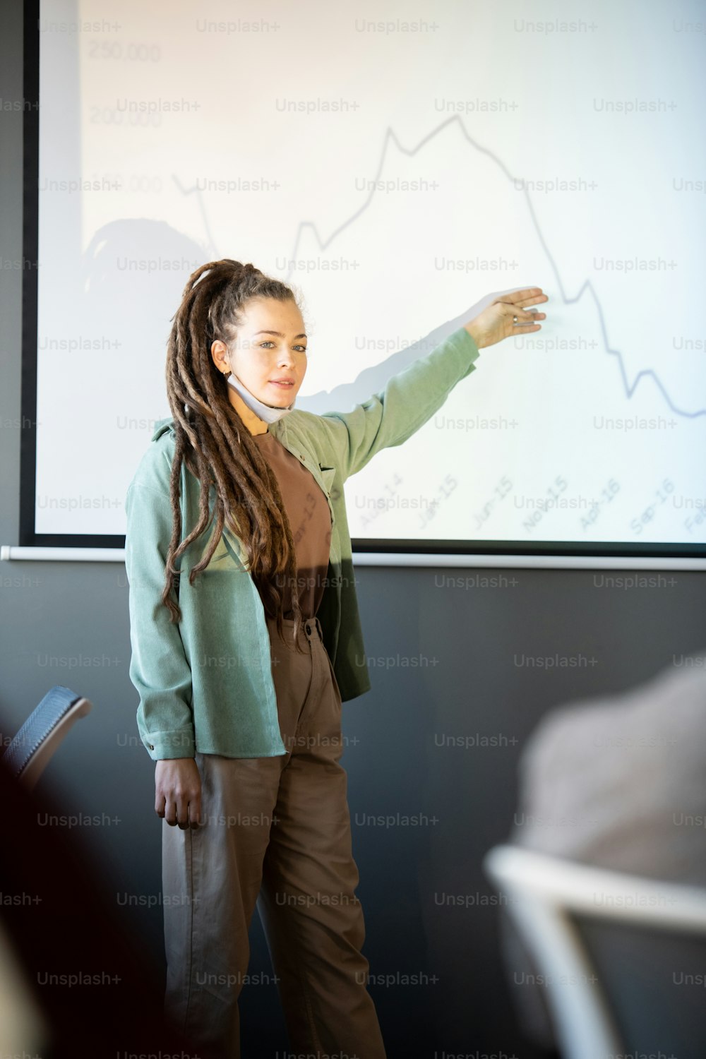 Giovane business coach femminile fiducioso in abbigliamento casual che spiega il grafico sulla lavagna durante la presentazione alla conferenza per economisti