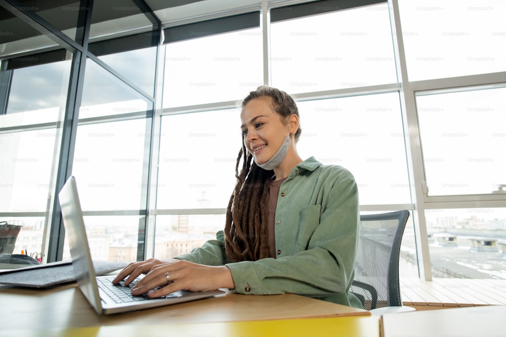 Giovane donna d'affari contemporanea felice in abbigliamento casual che utilizza il computer portatile mentre è seduta sul posto di lavoro contro la grande finestra nell'ufficio open space