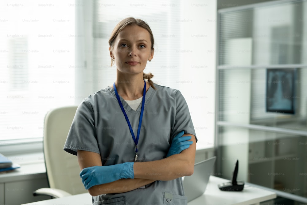 Retrato de una atractiva enfermera sonriente con guantes de látex azul con insignia en el cuello de pie con los brazos cruzados en la oficina
