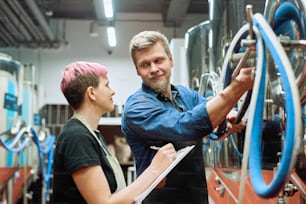 Young female worker of beer production factory making notes and looking at male colleague while both standing by large processing machine