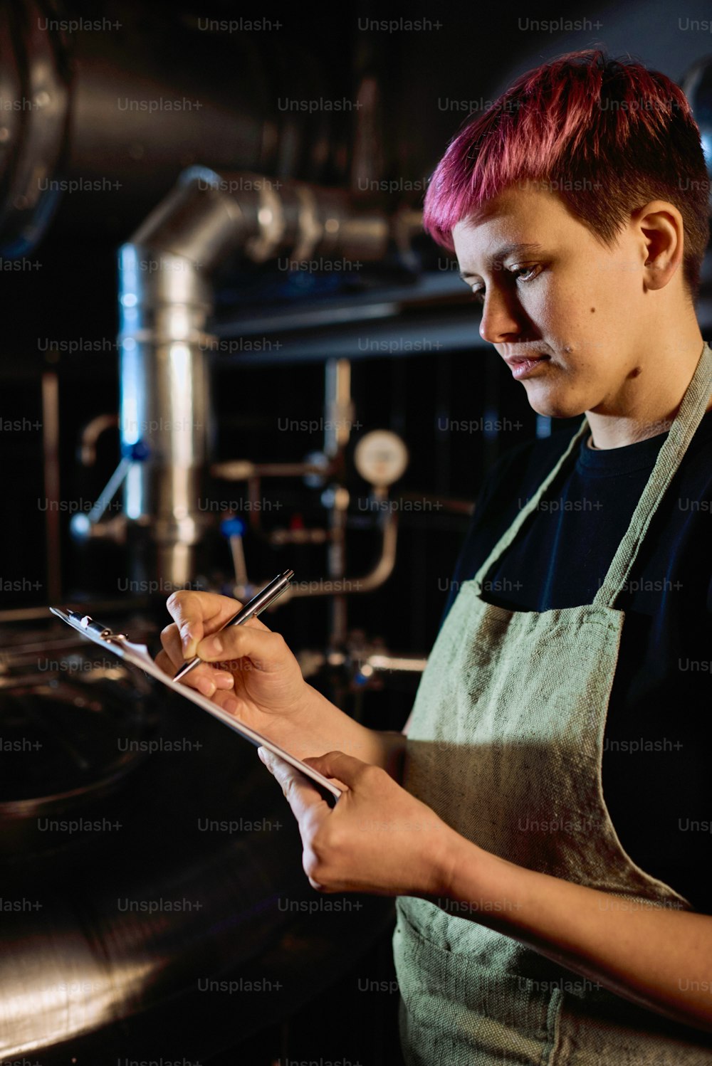 Young serious female brewer in apron pointing at document in clipboard while looking through list of stages of beer preparation at plant