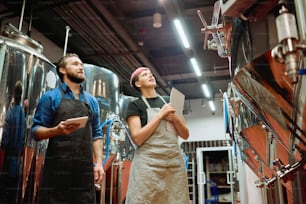 Two young successful workers of contemporary beer production factory looking at new equipment while discussing huge steel tanks