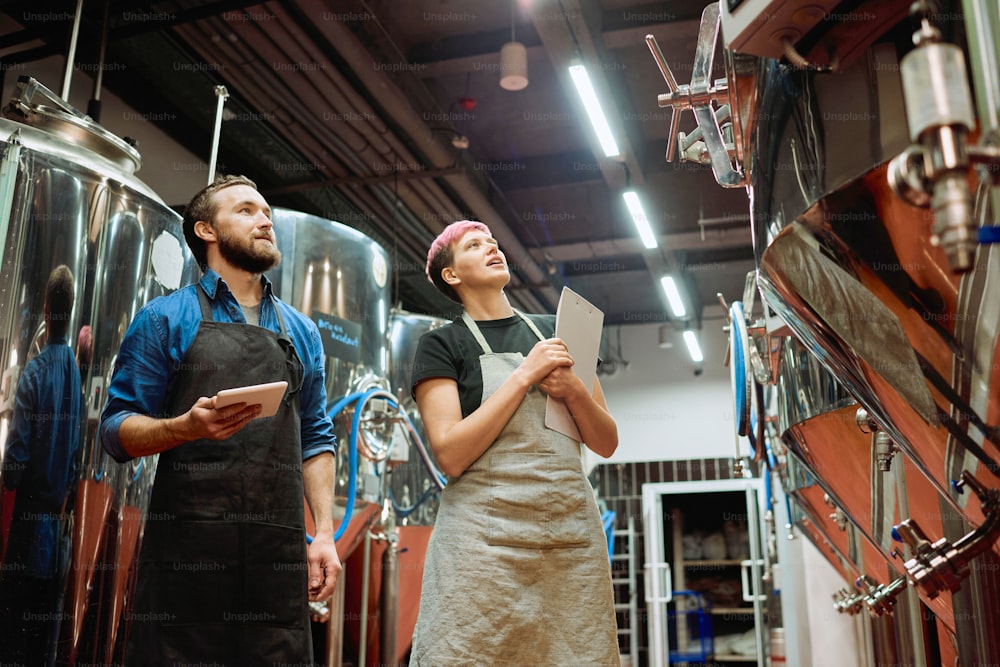 Dos jóvenes trabajadores exitosos de una fábrica de producción de cerveza contemporánea mirando nuevos equipos mientras discuten enormes tanques de acero