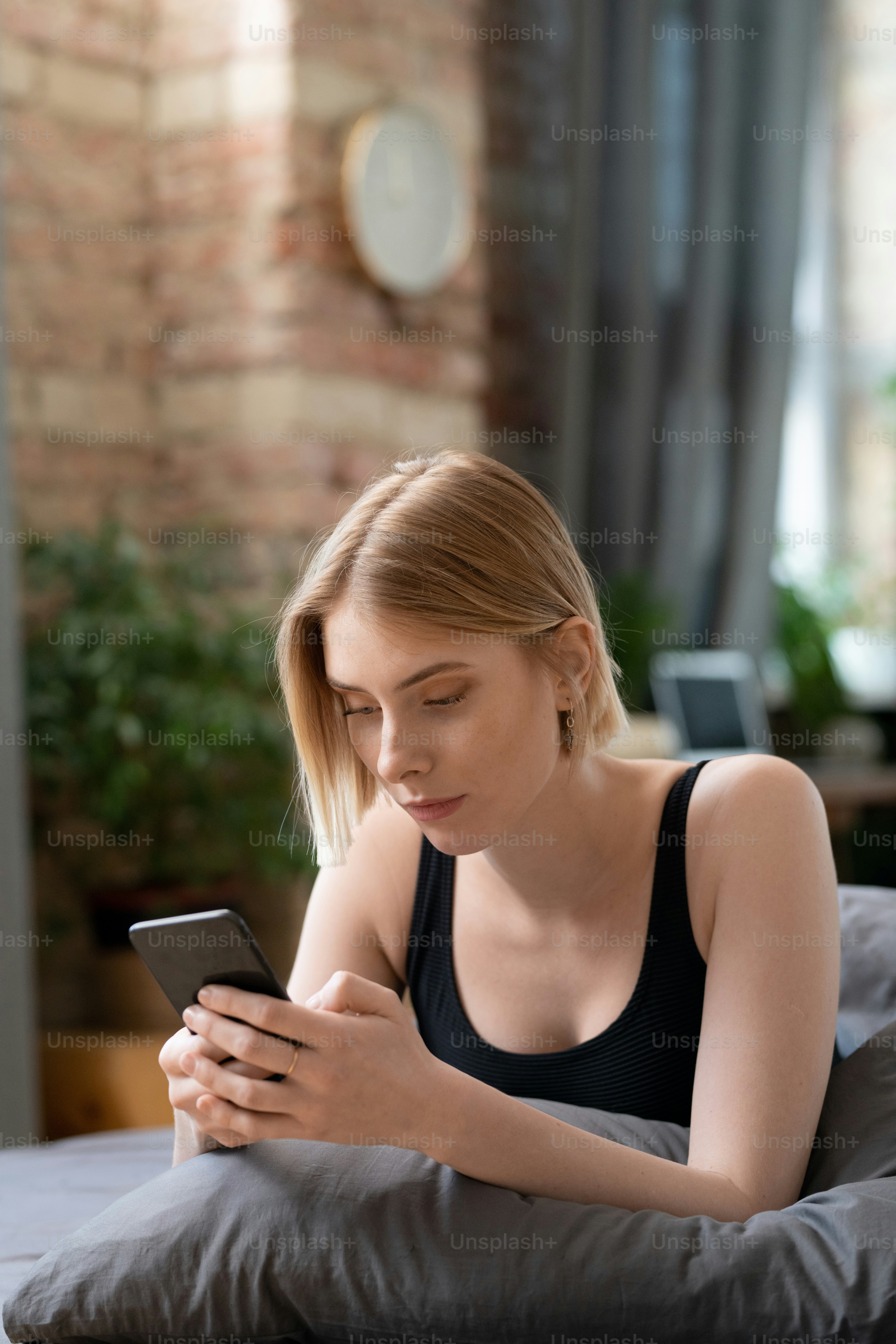 Young restful blond female with smartphone texting or scrolling through online news while relaxing on bed in bedroom on Sunday morning