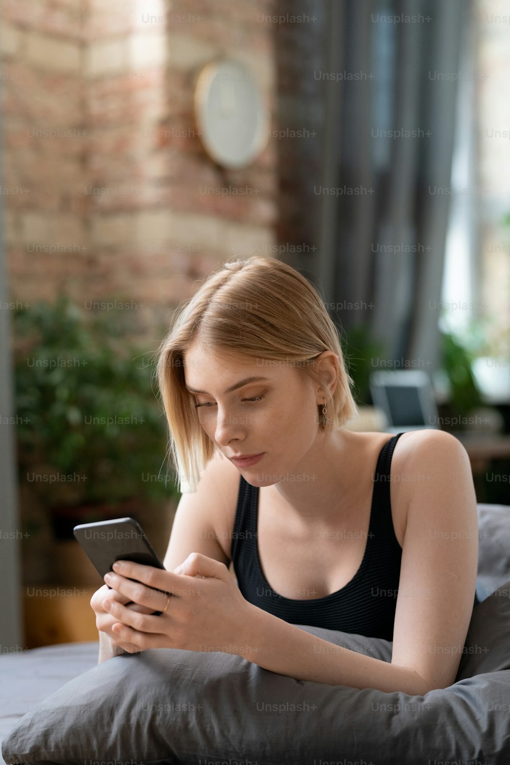 Jeune femme blonde reposante avec smartphone qui envoie des SMS ou fait défiler les nouvelles en ligne tout en se relaxant sur le lit dans la chambre le dimanche matin