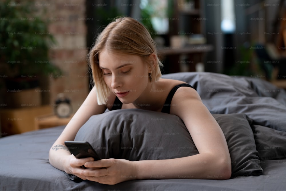 Mujer rubia joven con teléfono inteligente desplazándose por las noticias en línea o navegando en las redes sociales por la mañana mientras se relaja en la cama