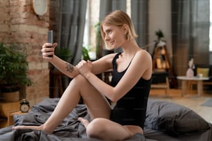 Feliz joven rubia con leotardo negro haciendo selfie o grabando un nuevo video para el blog en un teléfono inteligente mientras está sentada en la cama después de dormir