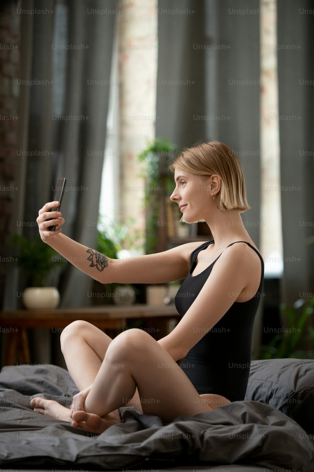 Mujer joven feliz con leotardo negro haciendo selfie o grabando un nuevo video para el blog en el teléfono inteligente por la mañana mientras está sentada en la cama después de dormir
