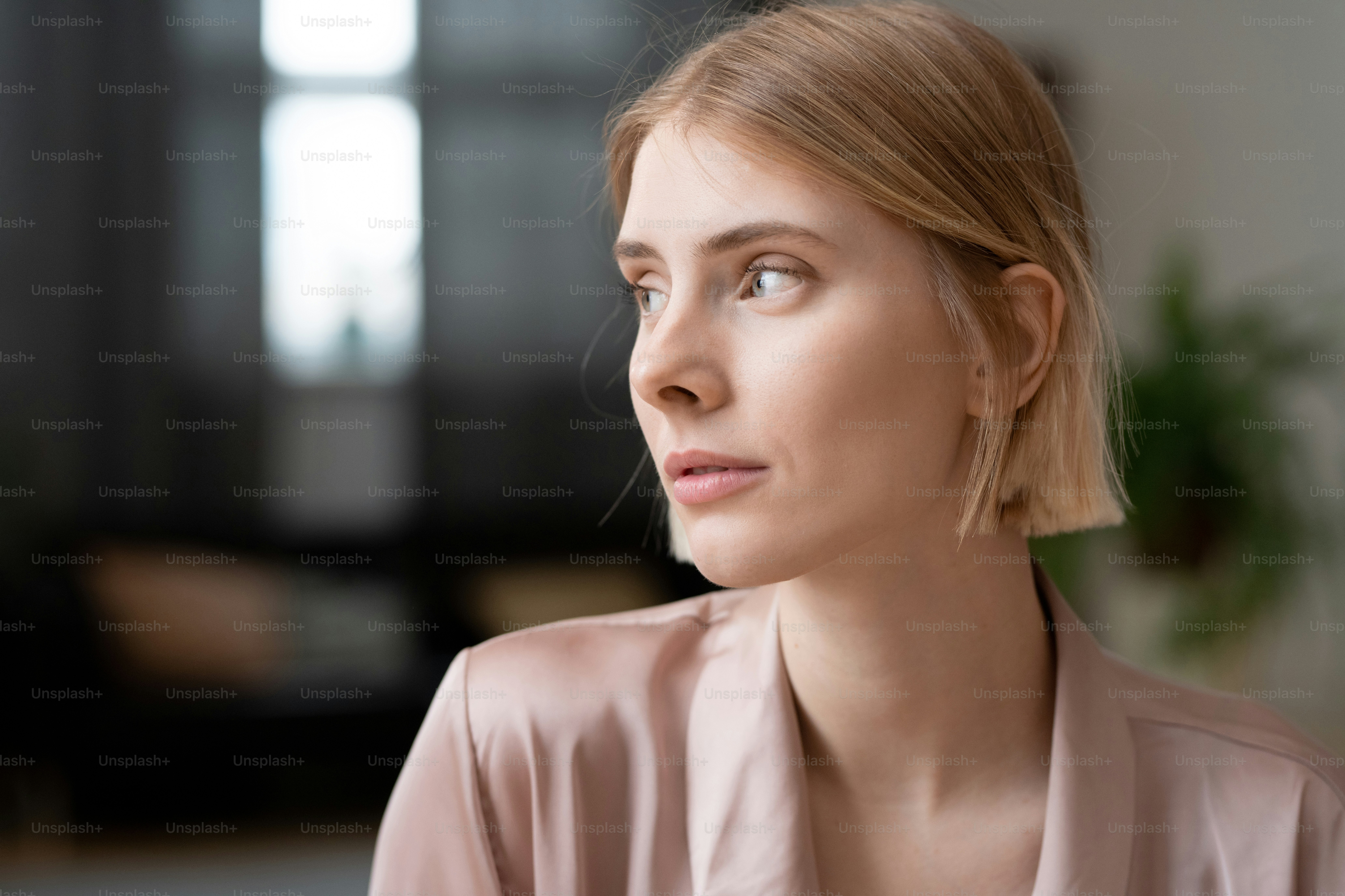 Young gorgeous blond female in beige silk robe or pajamas looking aside while thinking about forthcoming weekend in front of camera