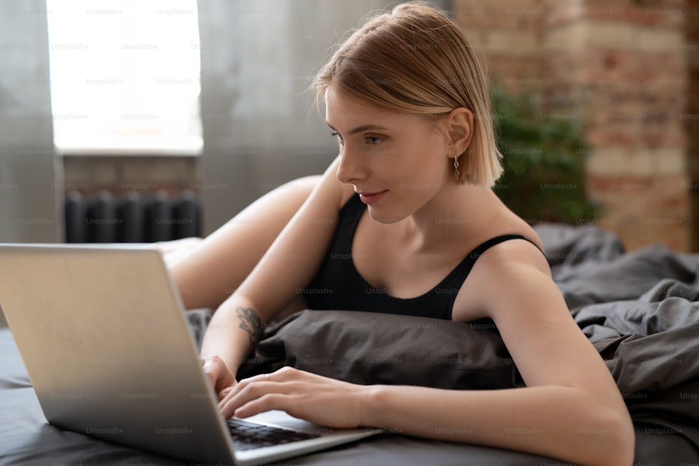 Mujer joven acostada en la cama y escribiendo en una computadora portátil que trabaja en línea en casa