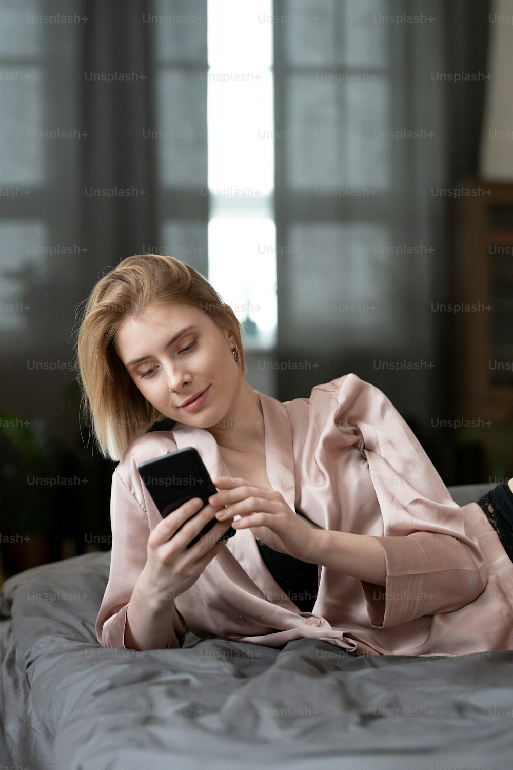 Bella donna in pigiama sdraiata sul suo letto e digitando un messaggio sul suo telefono cellulare nella stanza