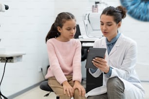 Young female ophthalmologist in whitecoat pointing at tablet display while showing schoolgirl result of check-up of her eyesight in clinics