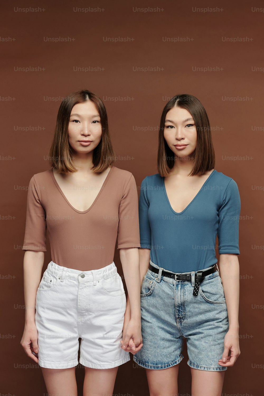 캐주얼한 풀오버와 반바지를 입은 젊은 아름다운 쌍둥이 자매가 스튜디오의 갈색 배경에 카메라 앞에 가까이 서 있다