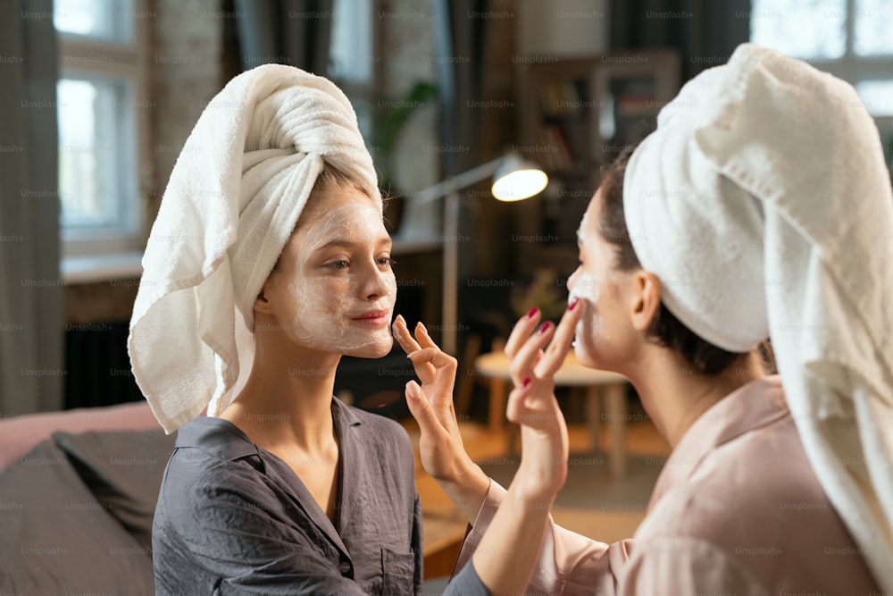 Two young women in silk pajamas and towels on heads applying clay masks on faces of one another while enjoying spa procedure at home