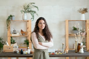 Happy young brunette woman crossing arms by chest while standing against workplace with ingredients for making natural cosmetic products