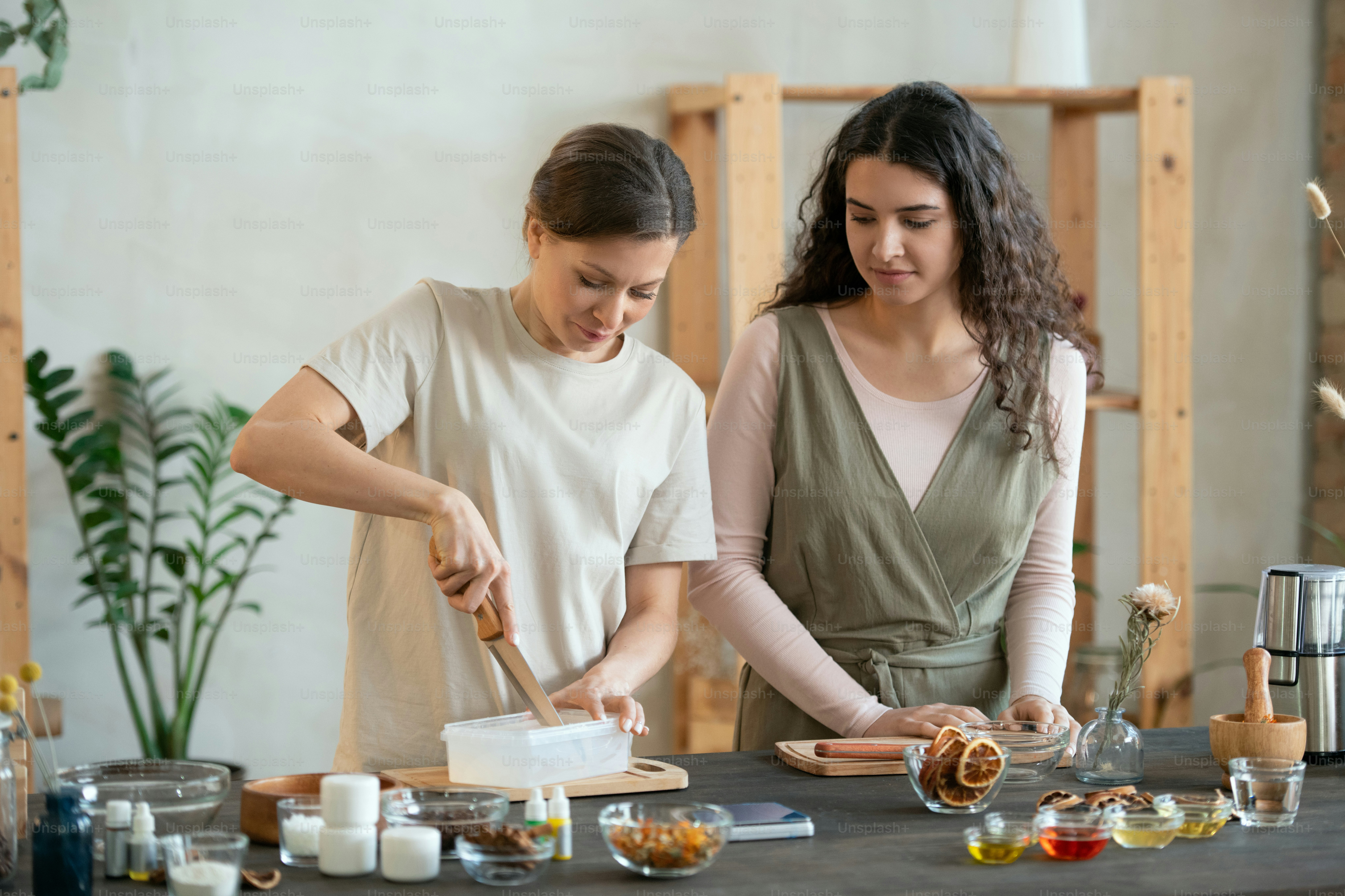 Zwei junge Frauen stehen am Tisch, während eine von ihnen harte Seifenmasse in Plastikbehältern schneidet, bevor sie Naturkosmetikprodukte herstellt