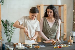 Two young females standing by table while one of them cutting hard soap mass in plastic container before making natural cosmetic products