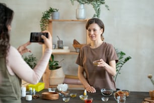 Joven mujer sonriente con un pequeño tazón que contiene aceite de esencia mirando la cámara del teléfono inteligente sostenida por su amiga mientras está de pie junto a la mesa