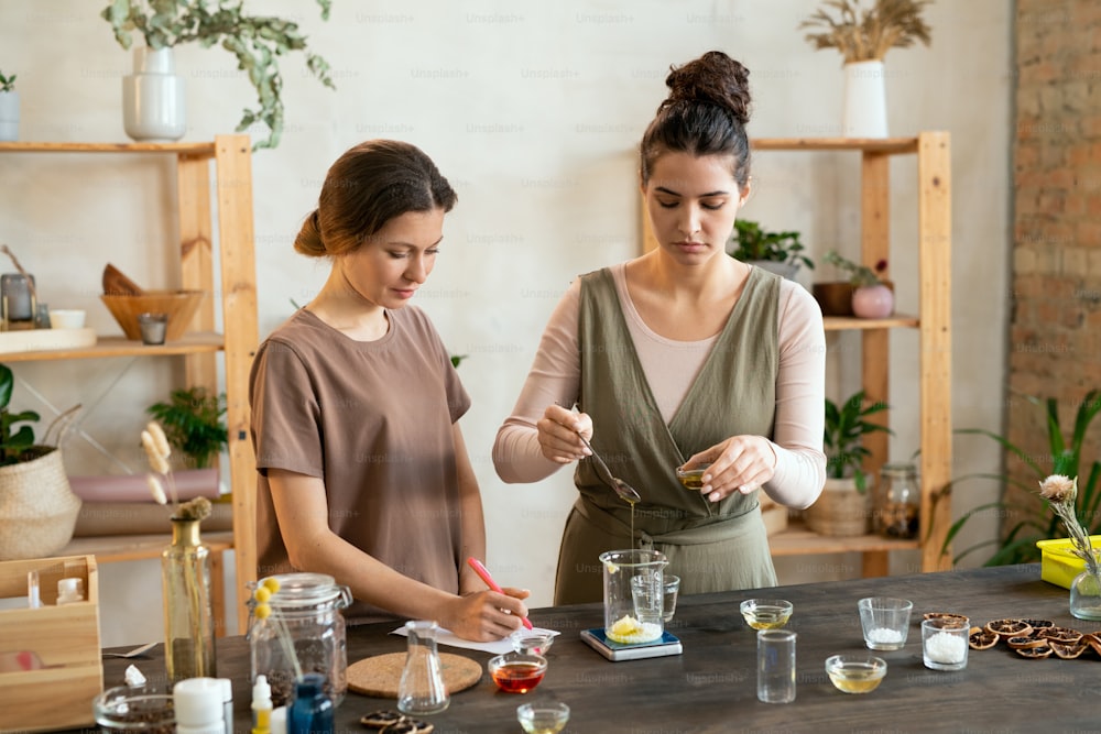 Eine von zwei jungen Frauen, die Essenzöl in Glaswaren mit gemahlener Seifenmasse geben, während sie eine Mischung für ein natürliches, handgefertigtes Kosmetikprodukt herstellen