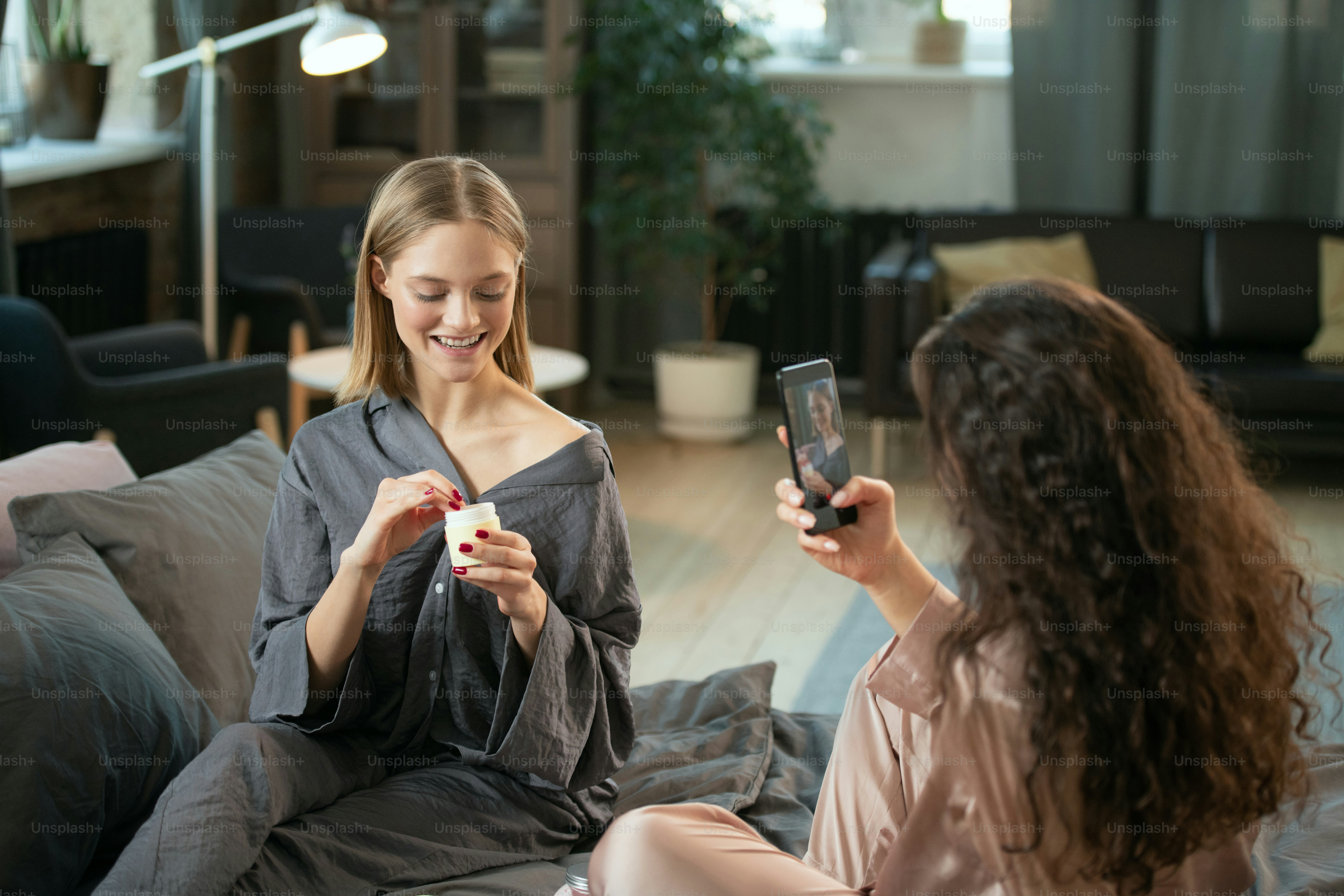 Cheerful girl applying natural handmade cosmetic product while sitting on bed in the morning, her friend with smartphone shooting her on camera