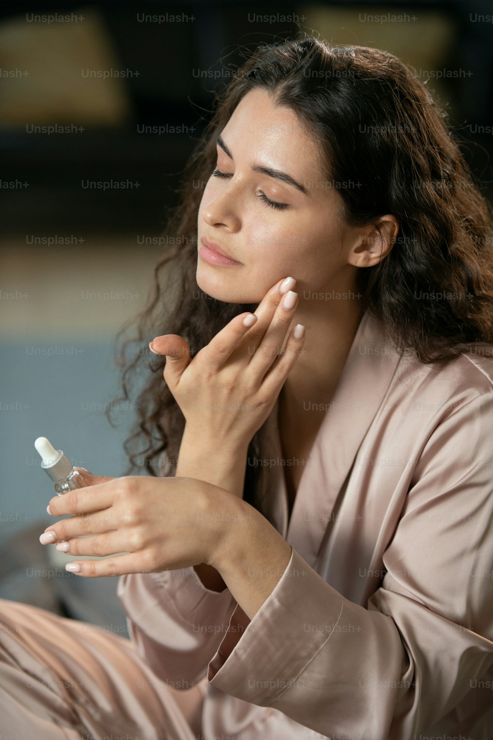 Young brunette pleased woman in silk pajamas applying natural cosmetic product on her face and enjoying it in the evening before sleep