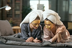 Dos mujeres jóvenes en pijama de seda, toallas en la cabeza y máscaras cosméticas doradas en las caras relajándose en la cama y desplazándose en el teléfono inteligente