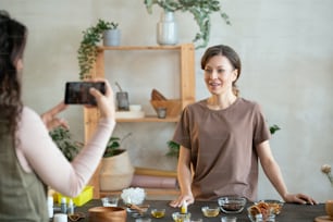 Young smiling woman with small bowl containing essence oil looking at smartphone camera held by her friend while standing by table