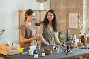 Manos de dos mujeres jóvenes cortando masa de jabón duro en cubos en tablas mientras están de pie junto a la mesa con ingredientes para hacer productos cosméticos.